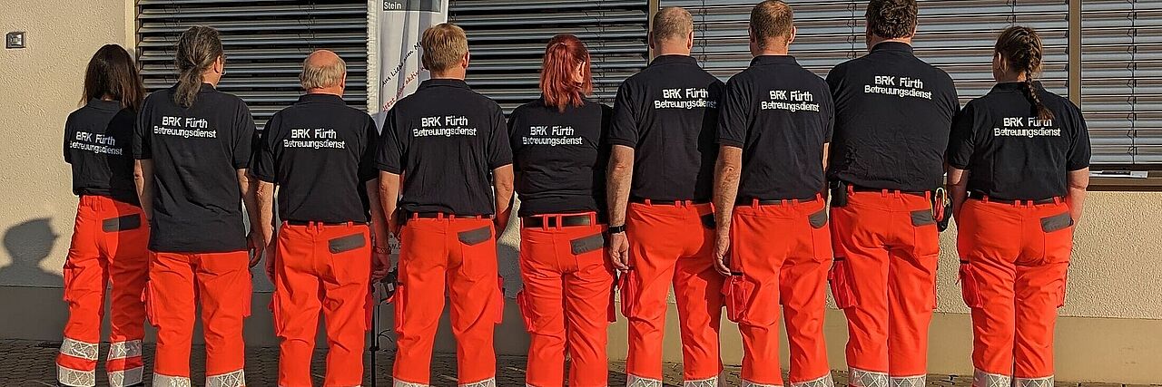 csm_betreuer2 mehrerer Helfer des Betreuungsdienstes stehen in Einsatzkleidung mit dem Rücken zum Fotografen vor einer großen Fensterfront